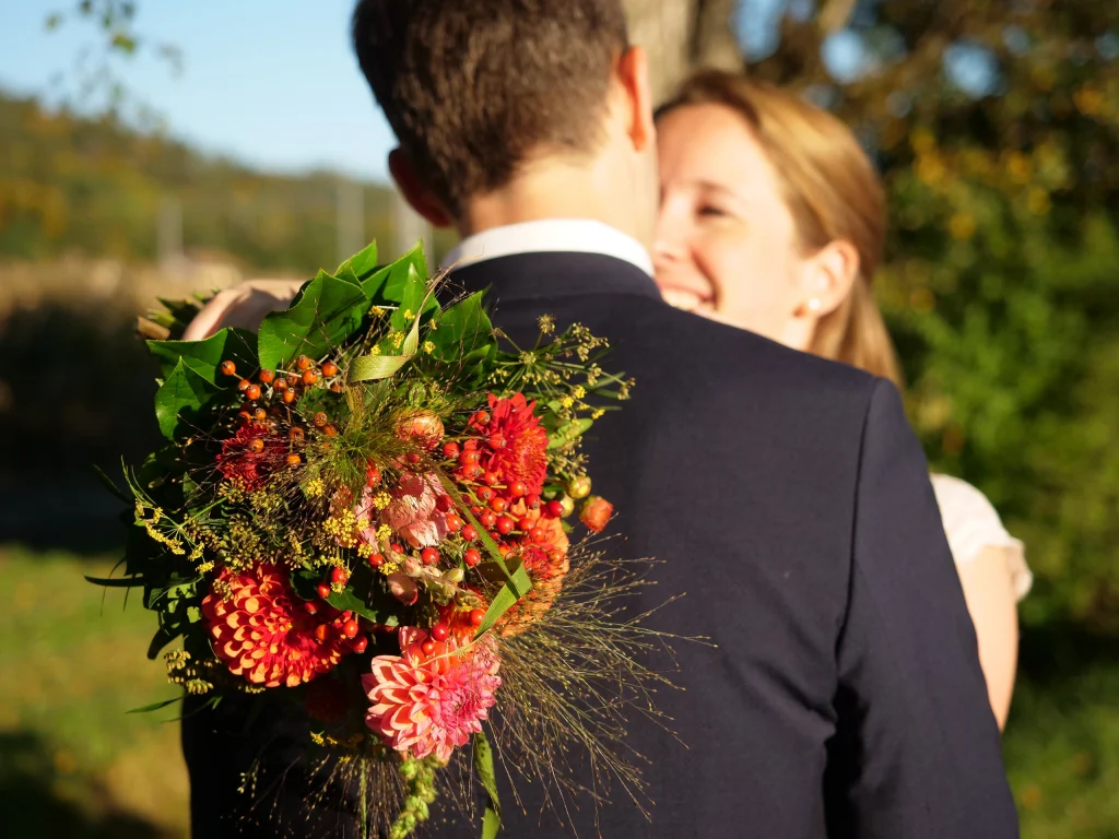 Ein Brautstrauss mit roten Blumen im Fokus. Er wird über die Schulter eines Mannes im Anzug gehalten, der der Kamera den Rücken zudreht, gegenüber von ihm ist unscharf eine Frau zu erkennen, die ihm ins Gesicht schaut