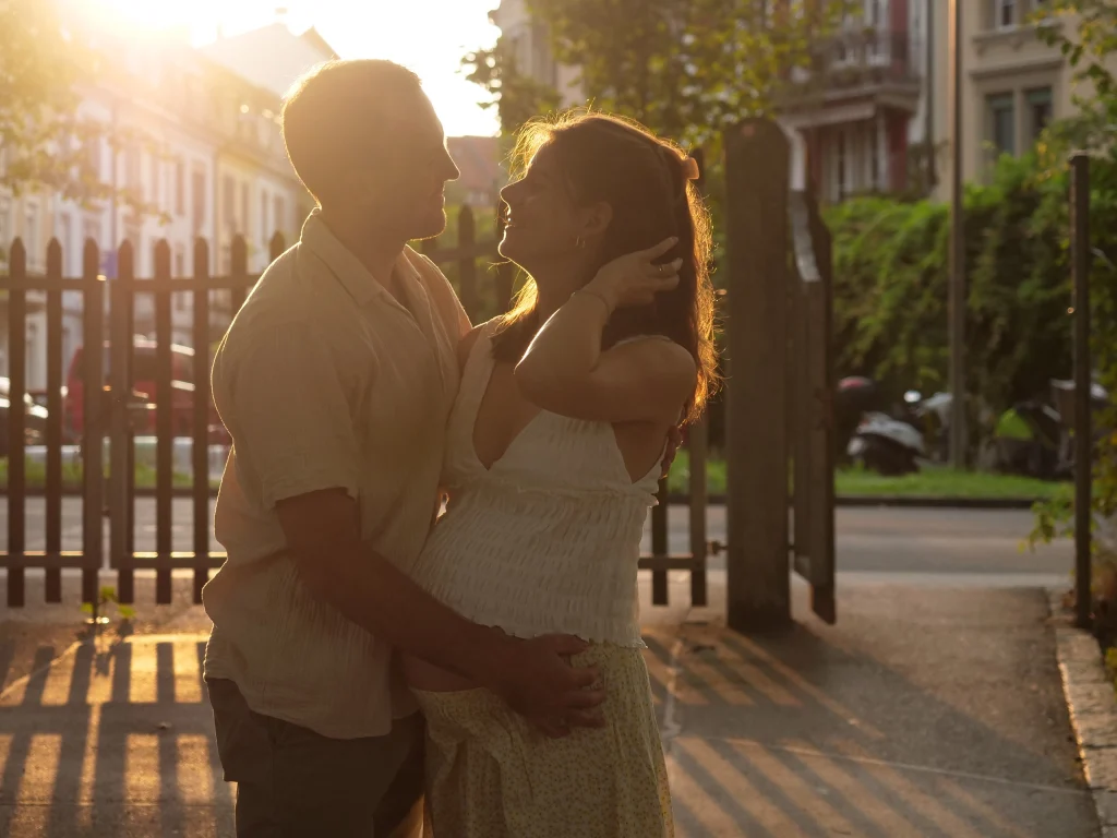 Im warmen Gegenlicht stehen ein Mann und eine Frau, die sich ins Gesicht schauen. Die Frau streicht sich mit einer Hand die Haare nach hinten, der Mann legt einen Arm um ihren schwangeren Bauch.